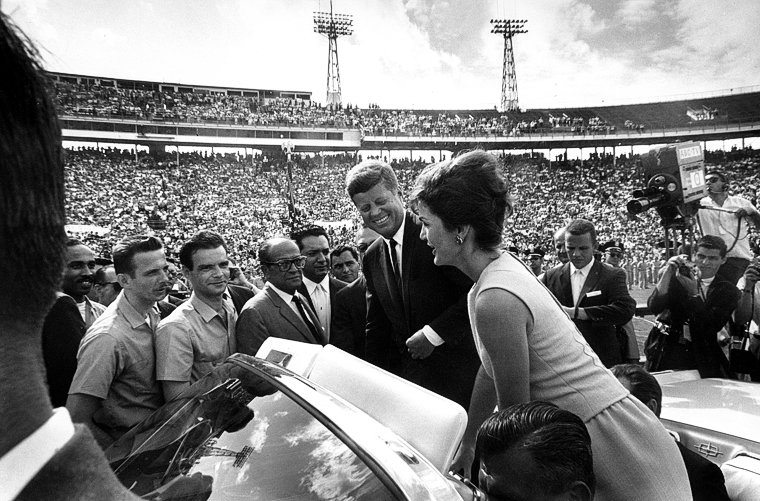 Jackie Kennedy speaking to gentlemen in stadium crowd alongside John F. Kennedy